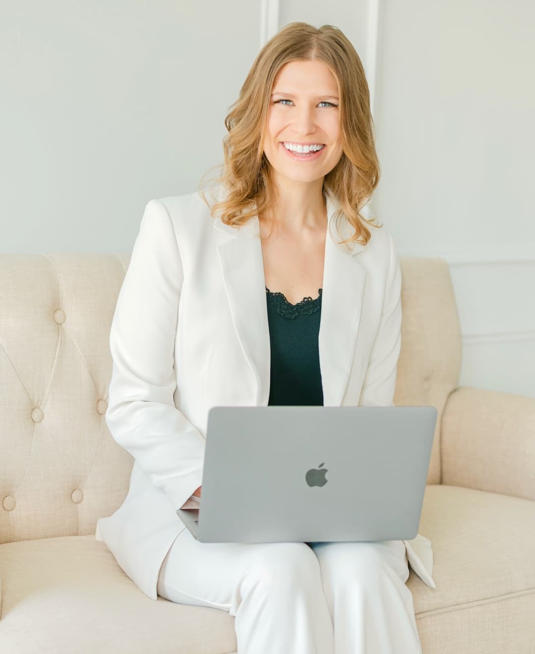 Christine Cornellier in a white suit, seated on a cream sofa with a laptop